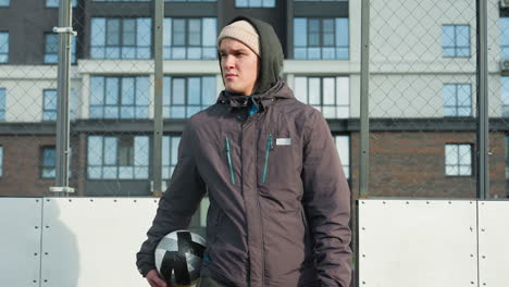 coach in hoodie and beanie holding ball in right hand walks along football pitch with goal post in background, tall office building in background, and sunlight casting shadow on him