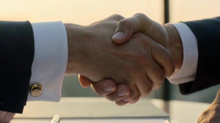 Mani di uomini che ispezionano, posizionano un dispositivo e una penna su documenti in ufficio, con gemelli, sigillando un accordo con una stretta di mano.