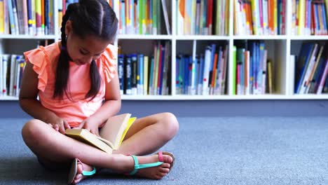 schoolgirl sitting on floor and reading a book in library at school