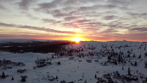 Breathtaking-aerial-view-of-a-sunset-casting-a-warm-glow-over-the-snowy-landscape-of-Trysil,-Norway,-with-scattered-trees-and-cabins