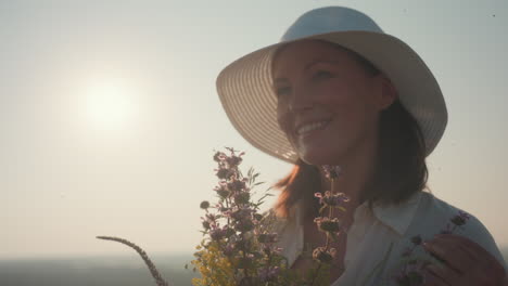young woman in white hat gently smells wildflowers in hand during sunset in quiet countryside, surrounded by soft sunlight and calm scenery