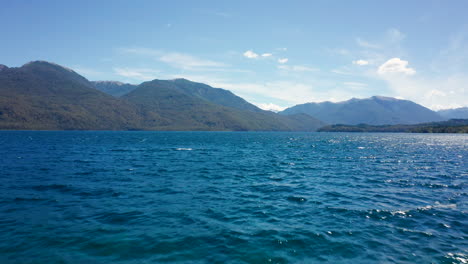 Forested-mountains-and-wavy-blue-water-in-Argentina,-low-forward-drone