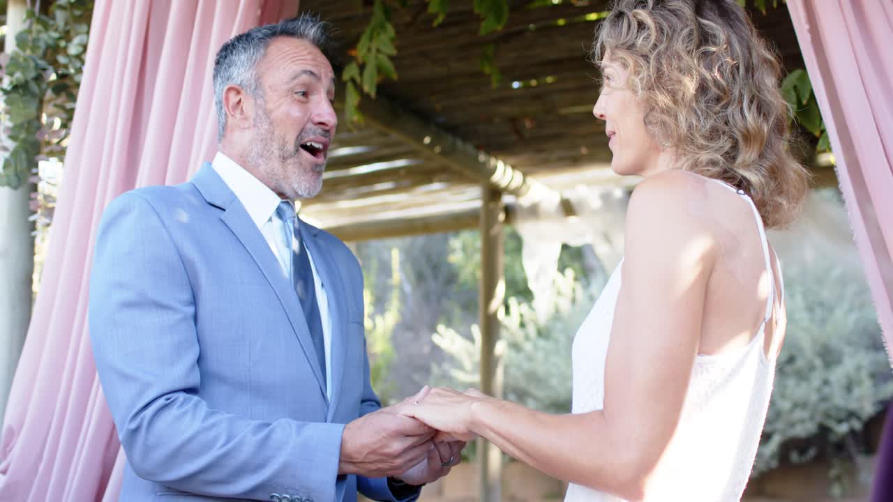Couple holding hands, man light-blue suit and woman white dress exchanging vows under pergola