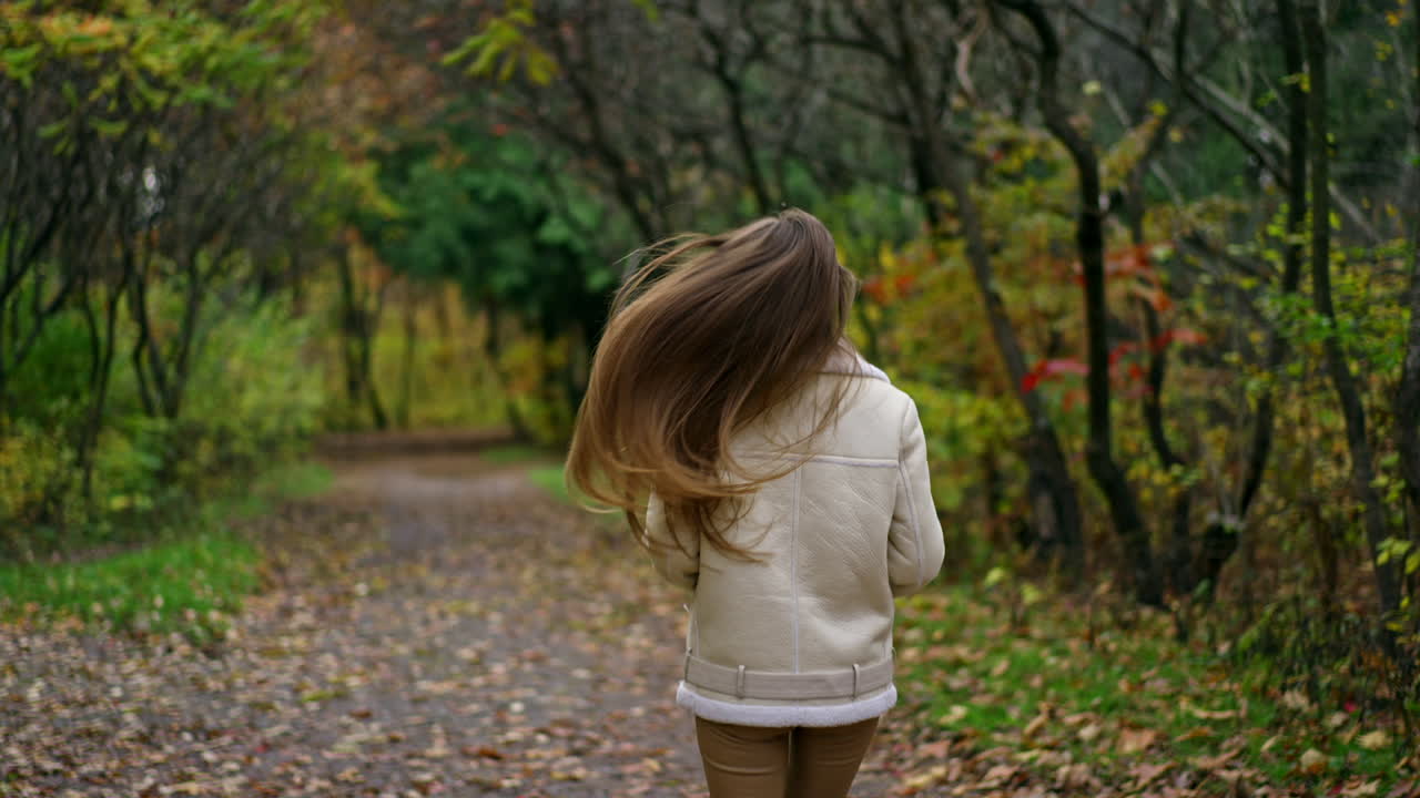 Attractive smiling girl running slowly in the autumn park. Brunette runs and her long hair moving beautifully. Blurred nature backdrop.