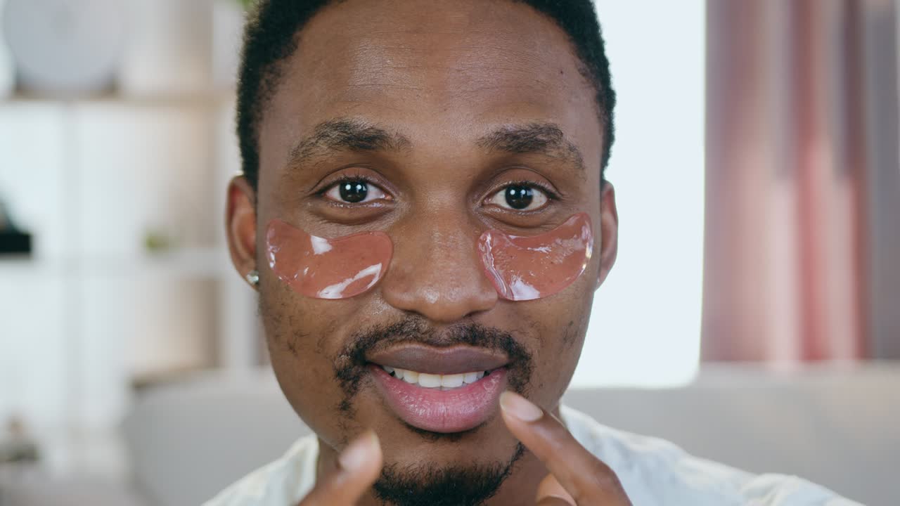 Face portrait of likable smiling caring young black-skinned guy which looking into camera with refreshing collagen eye-patches