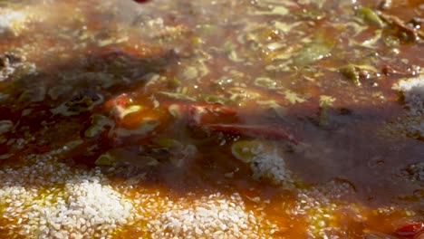 a steamy spanish paella dish is being garnished with rice from a pouch as it boils in the bright sunlight, slow motion, close up shot with a hand shadow above