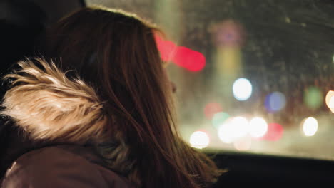 first-person view of a woman in a winter coat with a fur hood, inside a car at night without her head warmer. she is looking out thoughtfully,blurry city lights