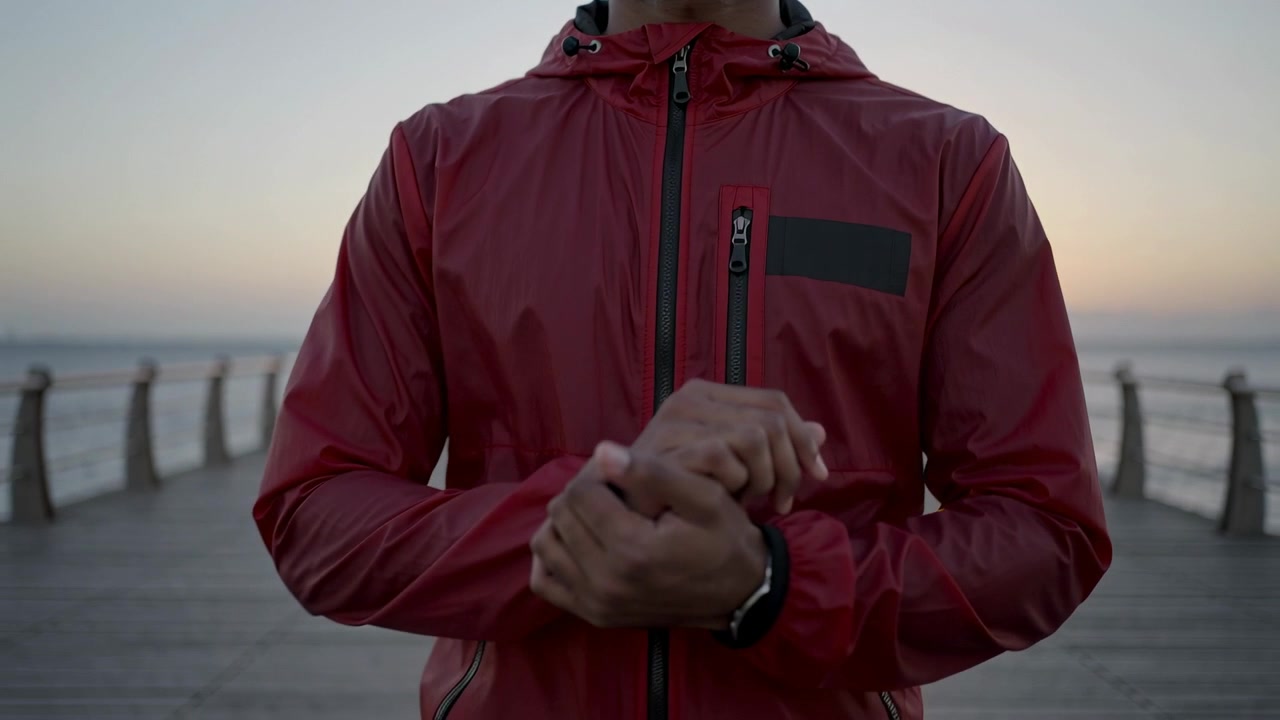 Noticing sunset man in red hooded jacket checking wristwatch on wooden pier, adjusting cuff