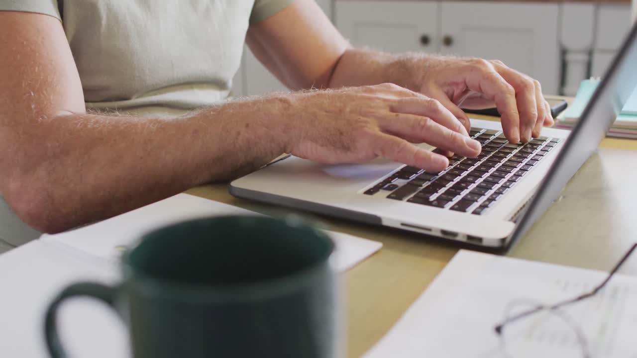 sección media de un hombre caucásico sentado en la mesa en la cocina, usando una computadora portátil