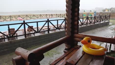 an inflatable duck lies on wooden table in gazebo at recreation center against the backdrop of pool covered with rain