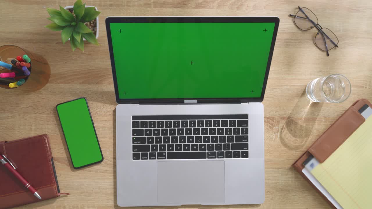 Top down view of a laptop computer with mock up green screen chromakey display on a wooden office desk next to notebook with pens, glasses, and a glass of water. Slow zoom out, close up