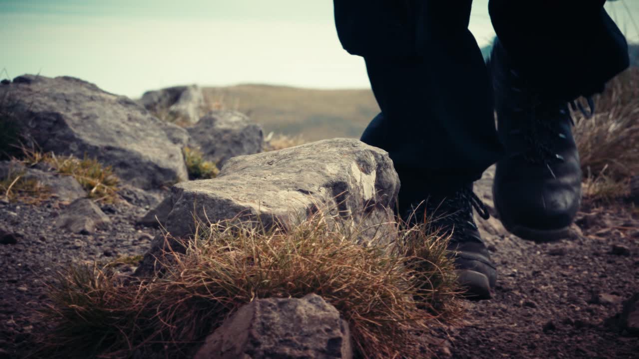 Nahaufnahme von Wanderstiefeln, die felsigen Boden mit trockenem Gras bedecken. Fokus auf die Textur der Steine und die Bewegung beim Wandern durch unwegsames Gelände.