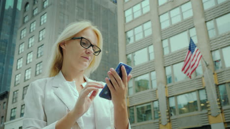 Businesswoman-Uses-Smartphone-On-The-Street-Of-New-York-On-The-Background-Of-Office-Building-With-Th