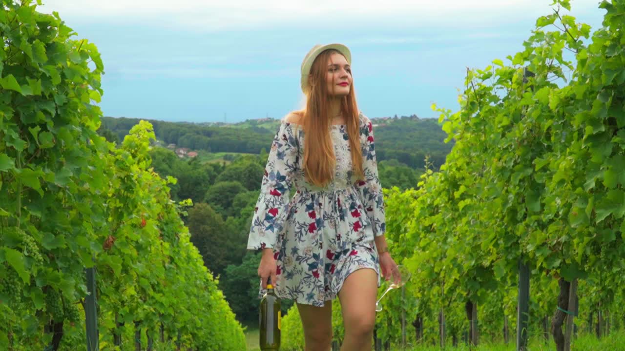Splendido filmato HD di una giovane donna bianca caucasica con cappello di lana, vestito e rossetto rosso che cammina tra i vigneti con una bottiglia di vino e un bicchiere in mano.