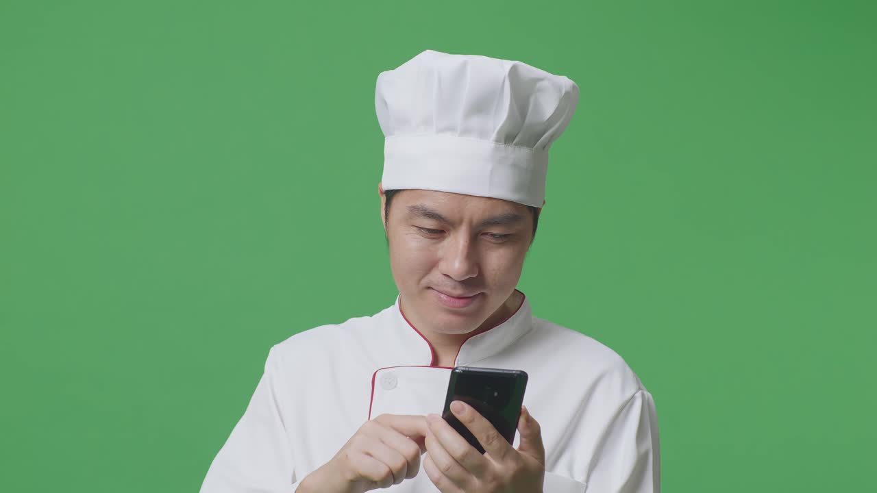 Close Up Of Asian Man Chef Smiling And Pointing To Smartphone In His Hand While Standing In The Green Screen Background Studio