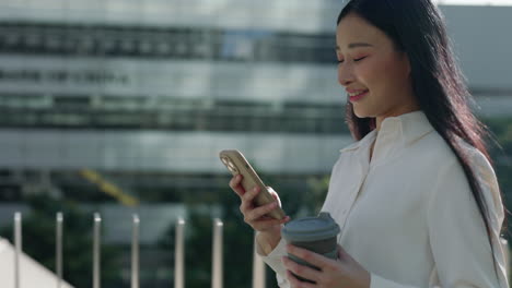 young asian woman smiling while using her smartphone and holding a coffee cup outdoors