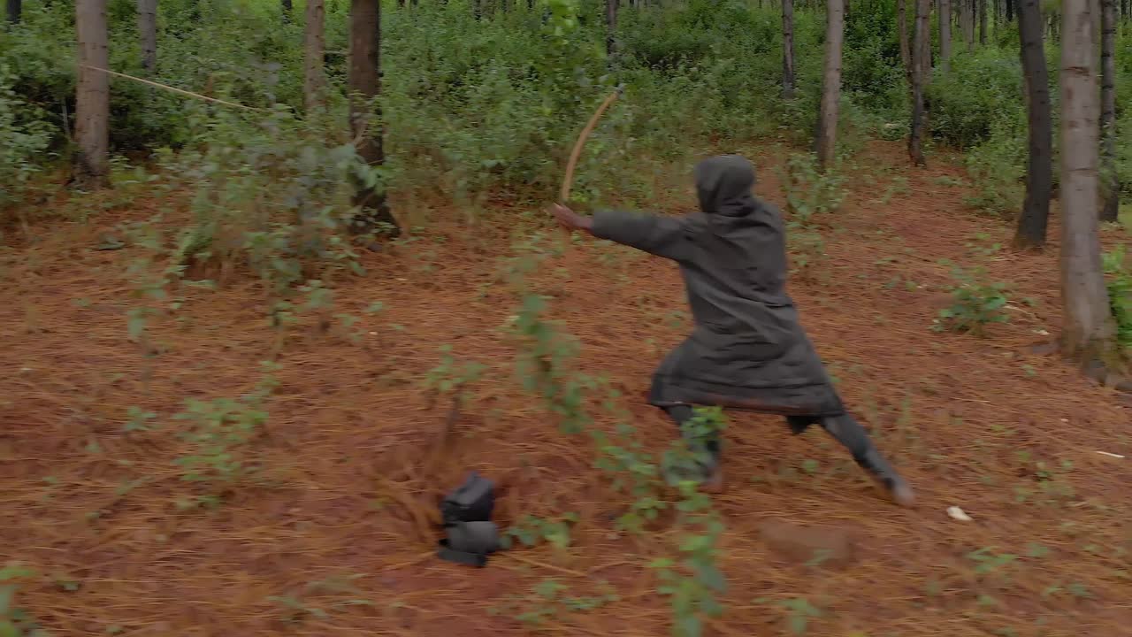 Aerial shot tracking an African hunter from the side as he walks through the bush takes aim and shoots a wooden bow an arrow