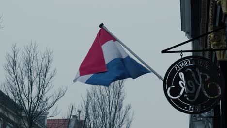 Toma-Estática-De-Una-Bandera-Holandesa-Ondeando-En-El-Viento-Fuera-De-Un-Café-En-Rotterdam