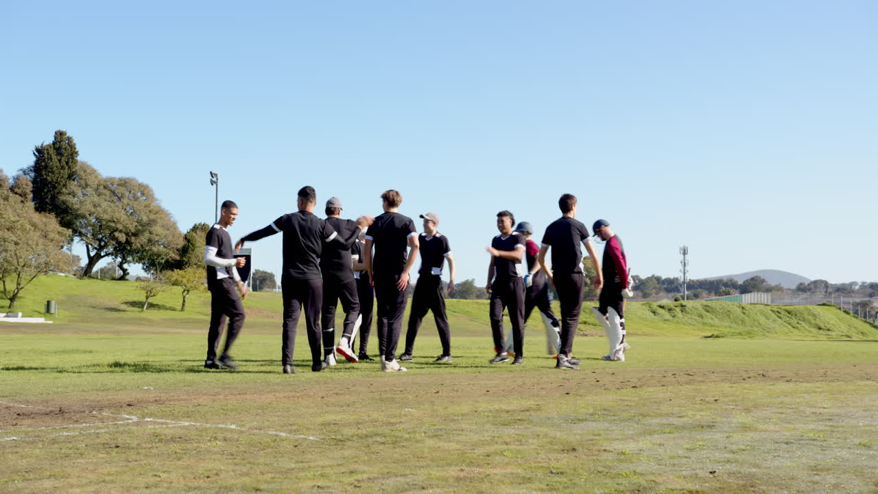 Two teams of multiracial male cricket players playing cricket, high fiving and huddling on pitch