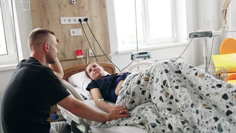man supporting his pregnant wife lying in hospital bed. husband hold his wife's hand talking to her.