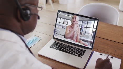 médico afroamericano que usa auriculares de teléfono tomando notas mientras hace una videollamada en una computadora portátil
