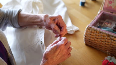 the wrinkled hands of an aging old woman untangling a knot in thread while repairing a button on a white dress shirt next to her sewing kit