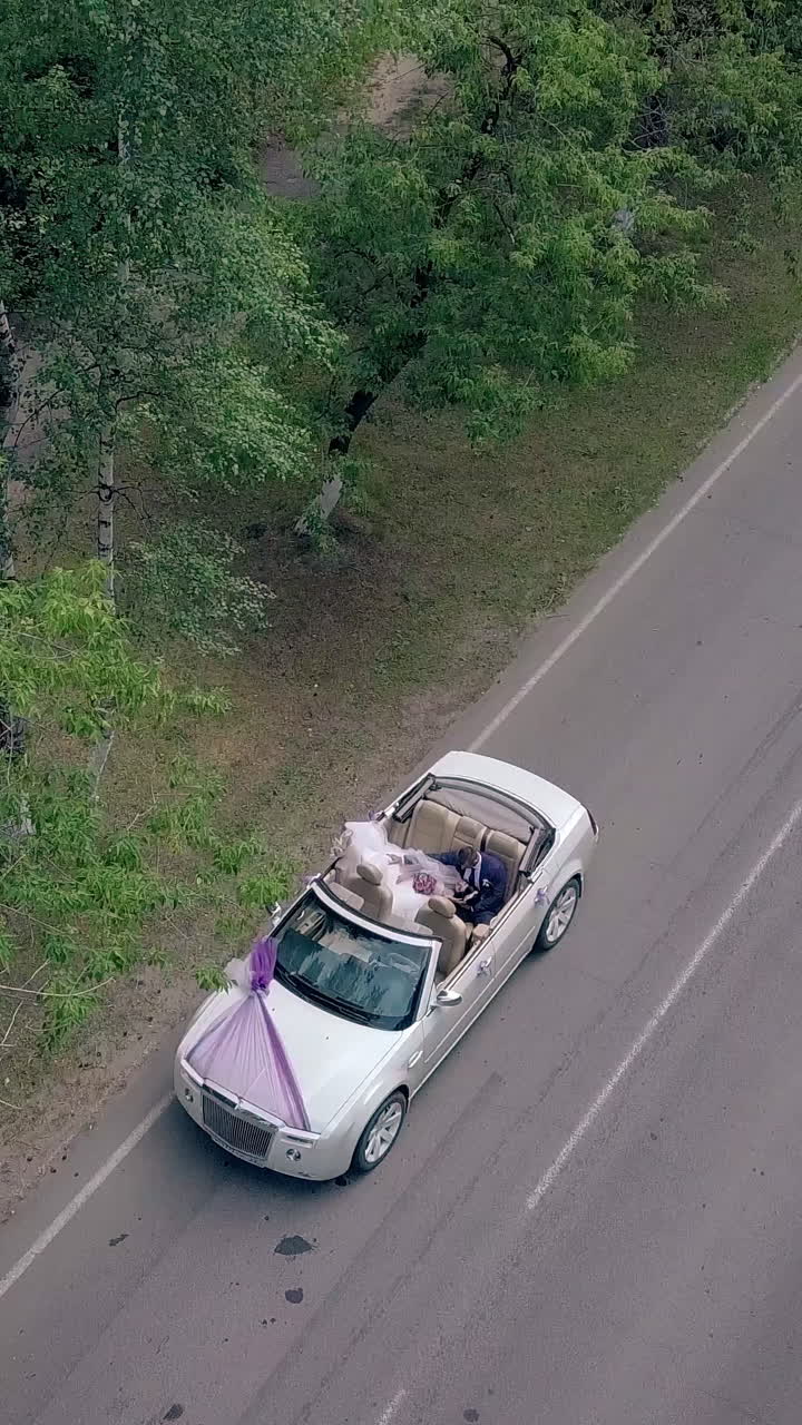 wonderful upper view couple sits in white celebratory wedding convertible on gray road among trees