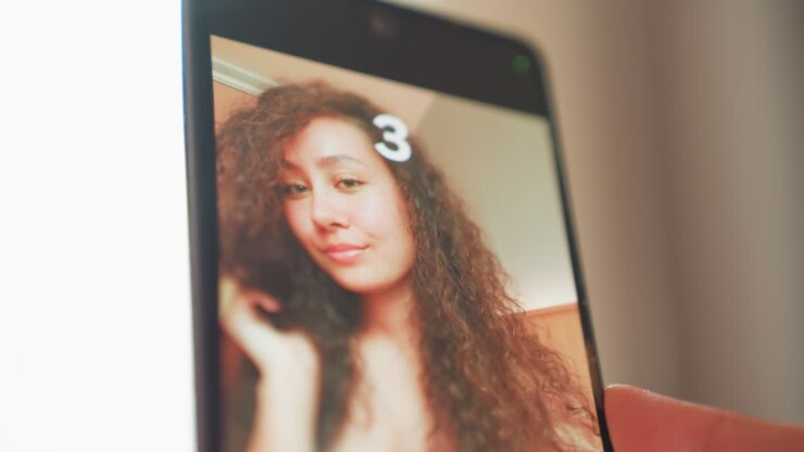 Selfie con smartphone che mostra una giovane donna che si sistema i capelli usando il timer per catturare un ritratto intimo: capelli ricci, illuminazione calda e soffusa, atmosfera da camera da letto, sorriso sottile, esperimenti con le angolazioni.