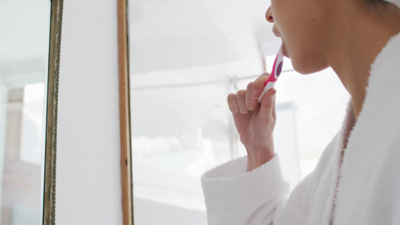 Woman in bathrobe brushing teeth while looking in the mirror