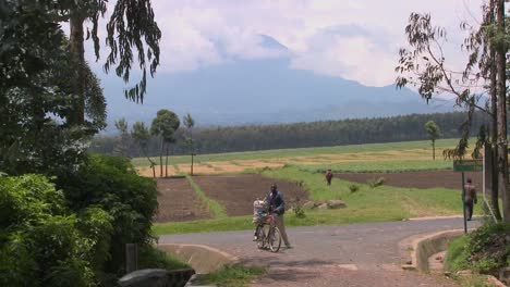 a man walks his bicycle down a rural road in rwanda with the virunga volcanos in the background