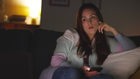 woman sitting on sofa at home at night streaming or watching movie or show on laptop and scrolling internet on phone 12