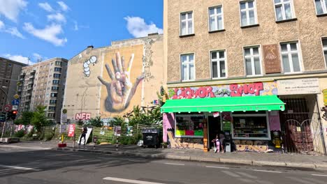 camera moves past a berlin city block with a large hand mural, liquor store, and bright storefronts under clear daylight, capturing urban street atmosphere