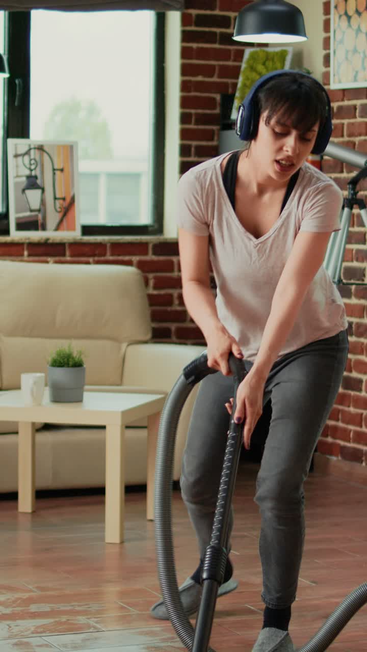 Vertical video: Female adult listening to music on headset and vacuuming floors at home