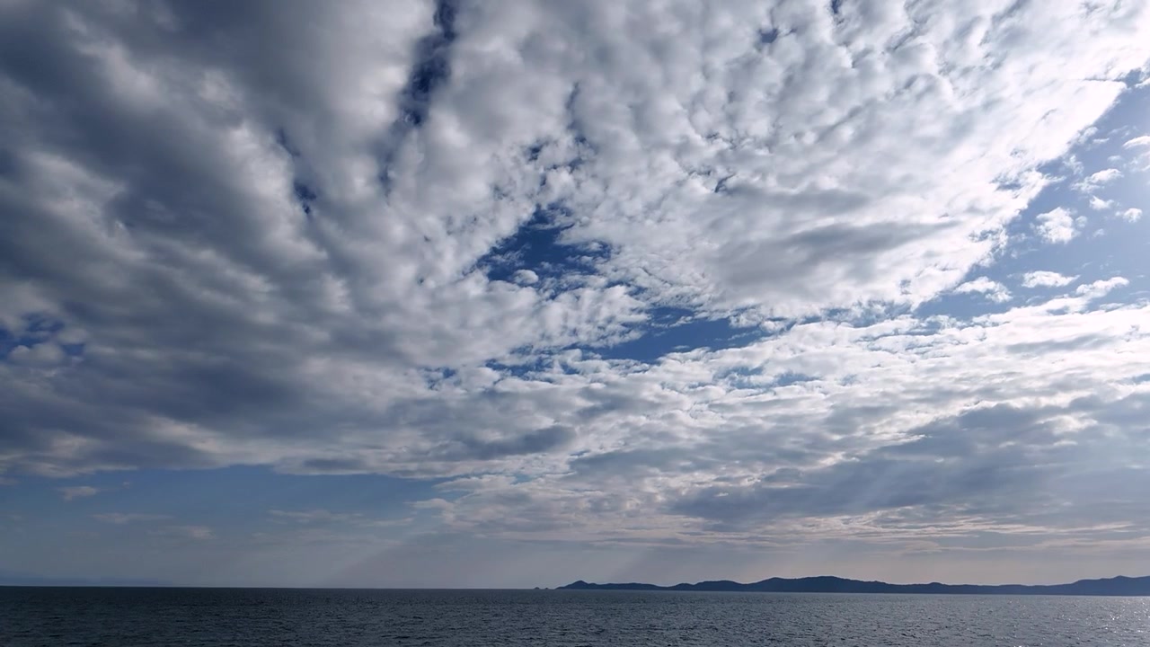 Awan melayang di langit di atas samudra pada sore hari yang cerah. Pemandangan ini menunjukkan permukaan air yang tenang memantulkan pola awan. Daratan yang jauh terlihat di cakrawala.