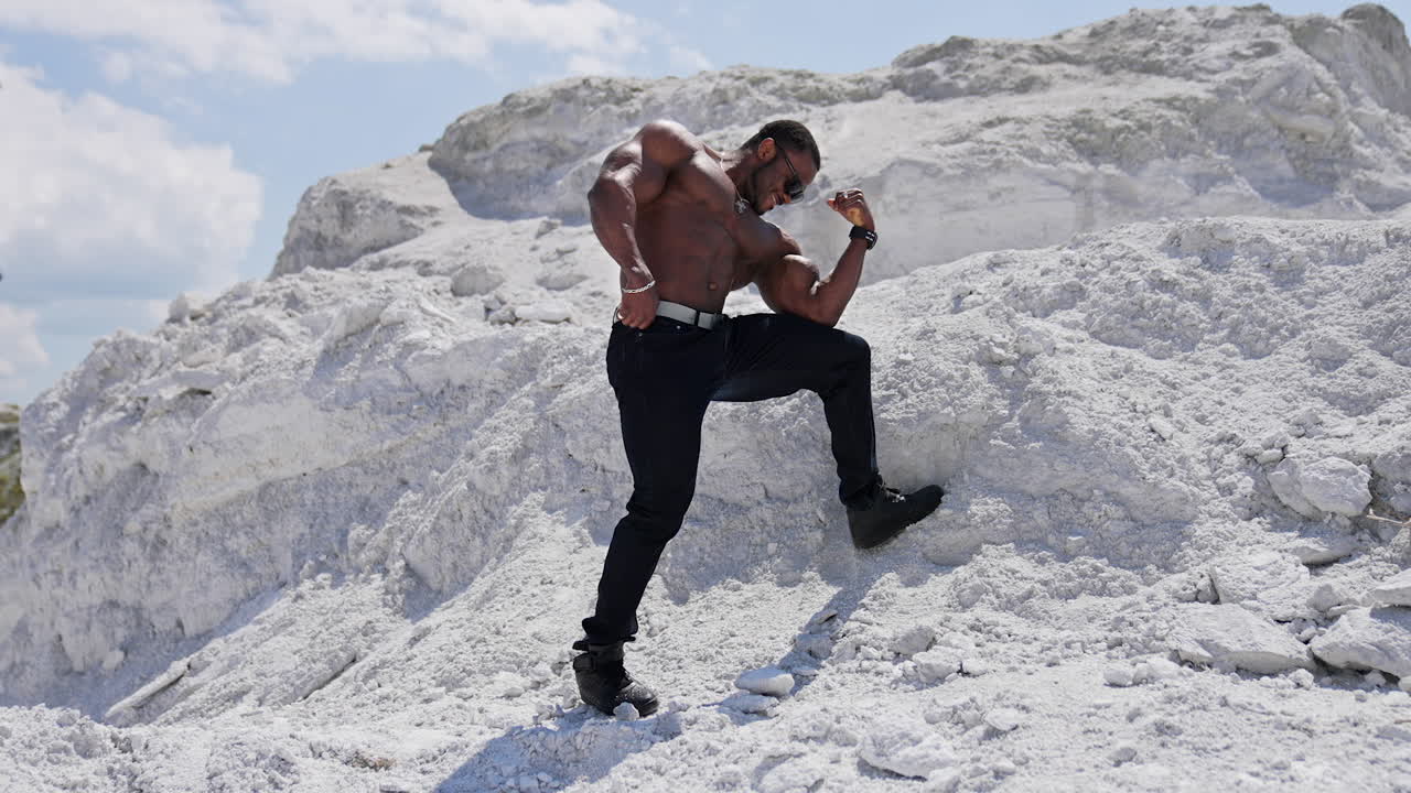 Handsome bodybuilder posing on camera. Sportive african american man in black glasses without shirt showing his strong muscles against white hill.