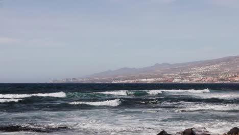 Olas-Rompiendo-Sobre-Rocas-Desde-El-Océano-Atlántico-Hasta-La-Costa-Con-Una-Ciudad-Al-Fondo,-Tenerife