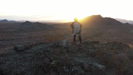 a lone man dressed as a spaghetti western hero stands atop a rugged mountain peak amidst a southwestern desert landscape as the sun sets in the distance. the camera makes a slow orbit around the man.