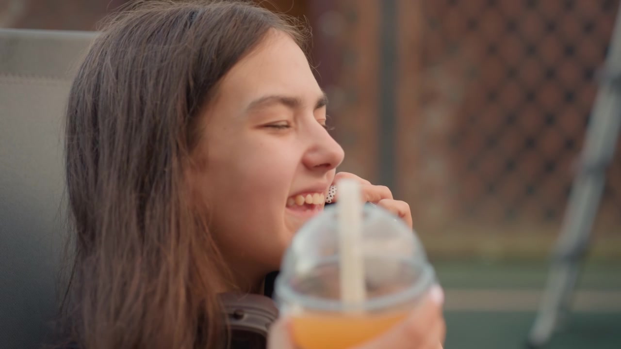 Young woman sits outdoors holding smartphone to her ear while wearing headphones around her neck. Woman holds drink with straw in other hand surrounded by fresh fruit slices on table. scene