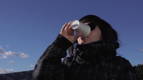 a girl in winter clothes, brown hair and sunglasses looking towards the camera and smiling