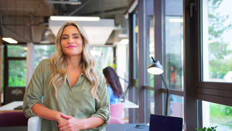 Portrait-Of-Smiling-Mature-Businesswoman-Sitting-On-Desk-In-Modern-Open-Plan-Office