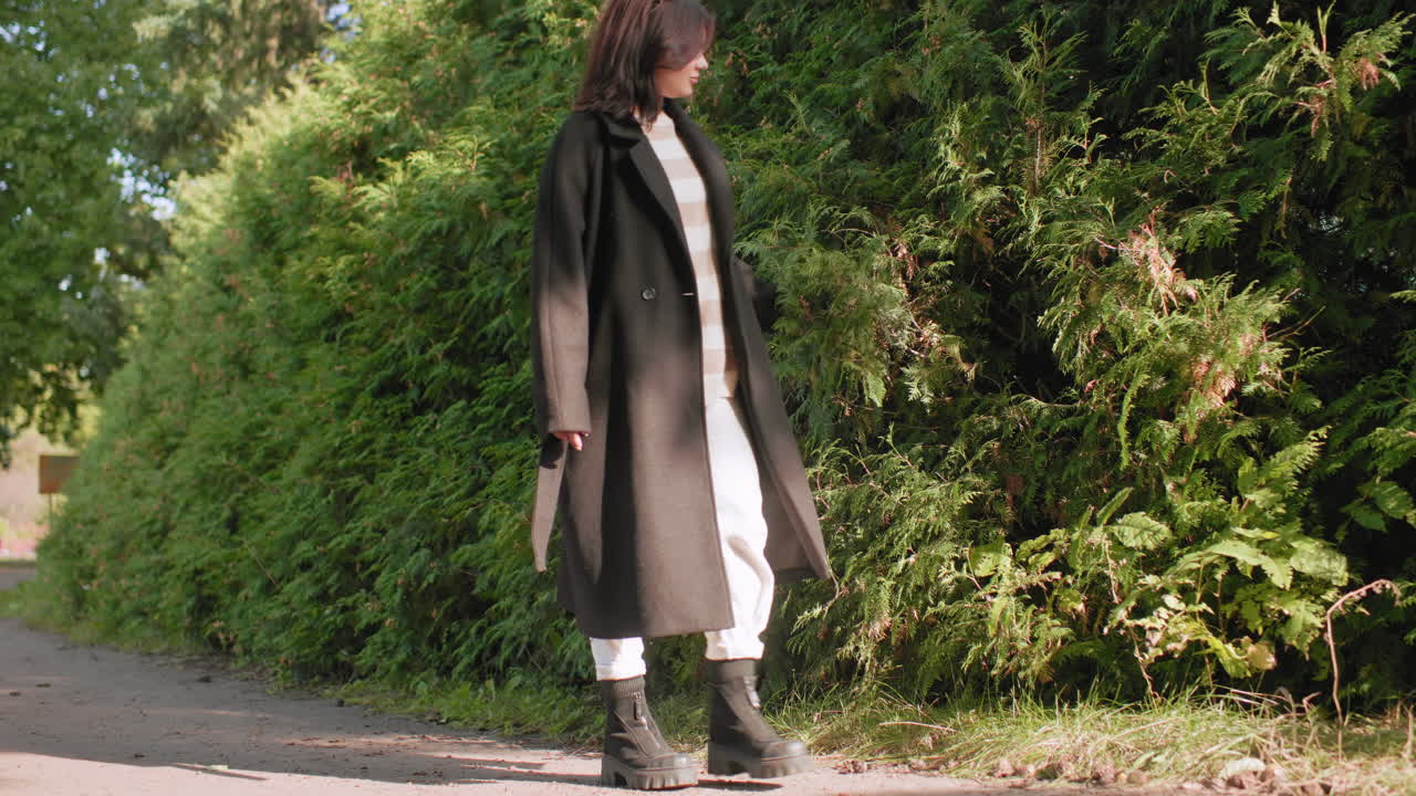 chica contemplativa caminando por un sendero del parque en un plano largo, su mano roza el follaje otoñal junto a un exuberante seto verde, lleva un abrigo y botas negras, pantalones blancos, ambiente tranquilo, enfoque en la naturaleza, luz suave del día