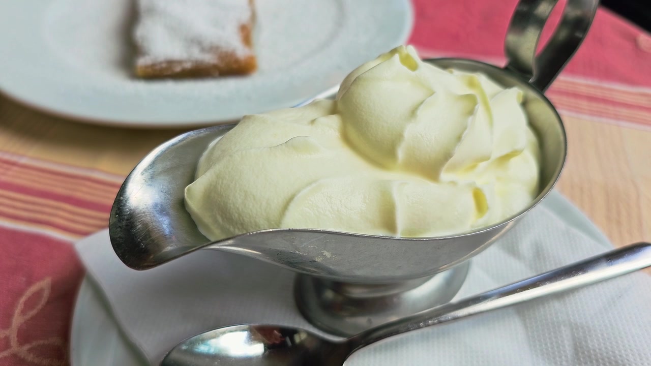 Morbido dessert alla crema di vaniglia in una ciotola d'argento con cucchiaio, pasticcino con zucchero a velo sfocato dietro, apparecchiatura informale da tavolino di caffè in un patio soleggiato di un ristorante europeo