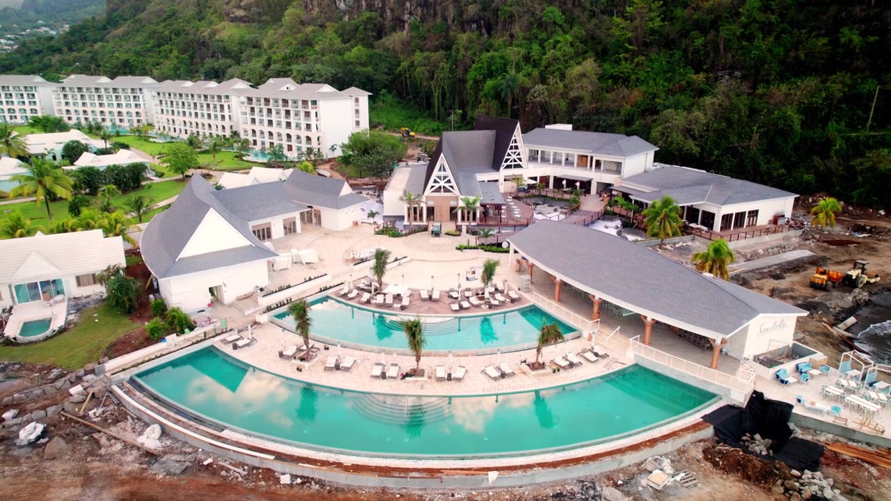 Una ripresa fluida con drone dall'alto che cattura il design a più livelli della piscina, gli edifici architettonici moderni e i terreni circostanti di un resort di lusso caraibico adagiato su un lussureggiante pendio montuoso.