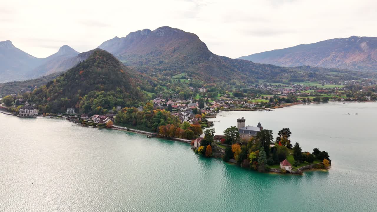 Dolly‑in drone shot over the peninsula of Duingt on Lake Annecy, moving towards Château de Duingt and the village backed by rounded Alpine hills and peaks in Haute‑Savoie, Auvergne‑Rhône‑Alpes, France