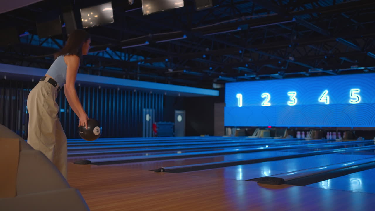 A brunette woman in bowling throws a ball in slow motion and knocks down skittles. Play bowling. Throw balls on the floor of the bowling club