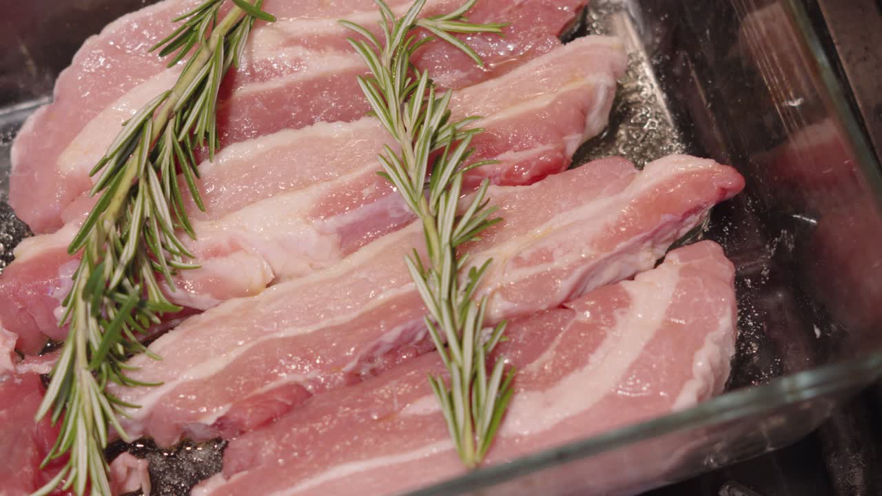 Fatias frescas de barriga de porco crua em travessa de vidro com sal e tempero de alecrim, prontas para assar no forno. Receita de jantar saborosa à base de carne. Cena de comida mostrando textura e ingredientes inteiros.