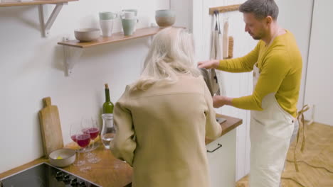 man washing the family dinner dishes at the sink in the kitchen while two mature women removing the plates from the table