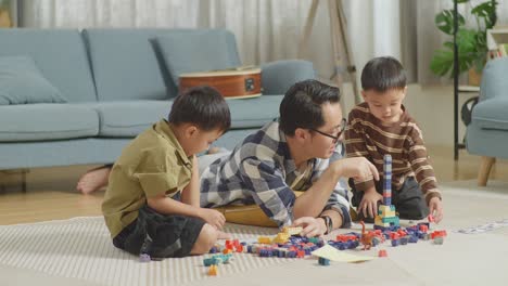 full body of asian father and sons playing the construction set colorful plastic toy brick at home. the kids assemble plastic building blocks sitting on a mat, a father lying on the floor talking to his sons