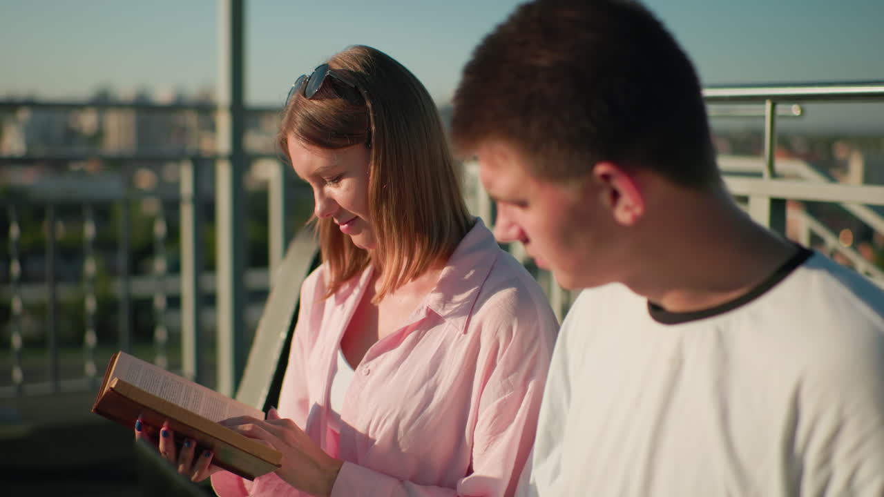 Friends seated outdoors studying as the lady explains something to the boy, who nods in agreement, the blurred background features greenery, urban buildings, and bright sunlight
