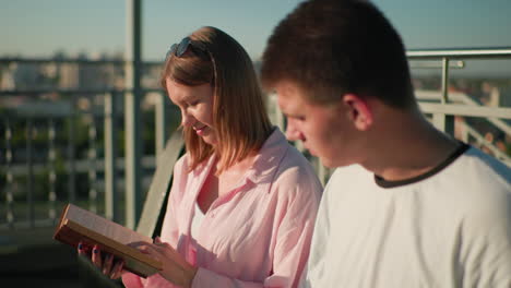 friends seated outdoors studying as the lady explains something to the boy, who nods in agreement, the blurred background features greenery, urban buildings, and bright sunlight